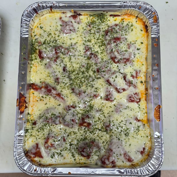 A rectangular aluminum foil pan filled with baked pasta, melted cheese, and red sauce, topped with dried herbs.