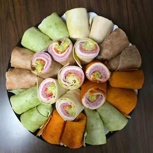 A platter of colorful wraps, some sliced to reveal ham, cheese, lettuce, and tomato fillings.