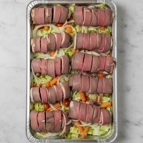 Sliced roast beef rolls with lettuce, onions, carrots, and olives in a foil tray.